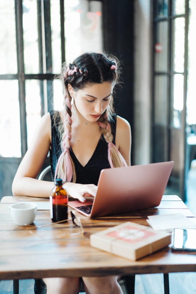 Young Woman Sitting In Coffee Shop At Wooden Table. On Table Is Laptop.